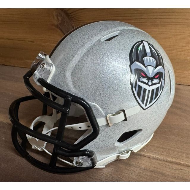 Michigan Arsenal Mini Helmet. Metallic silver shell with a black facemask. Dark knight-helmet face logo on the side with red eyes and a green accent stripe.