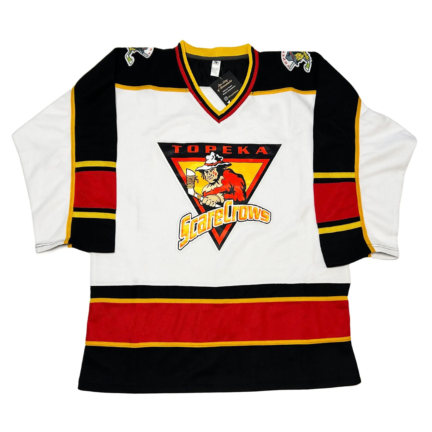 Topeka Scarecrows hockey jersey white. Black shoulders. Half white sleeves, half black, separated by a gold line. Thin gold, thin black, thick red, thin black, thin gold ban mid-sleeve. Black, and red ban separated by gold line at waist. Logo in center is upside down black triangle. TOPEKA in red above a scarecrow holding a hockey stick over a gold triangle outlined in red. Stylized SCARECROWS in gold arched across bottom of main Triangle. Royal Retros