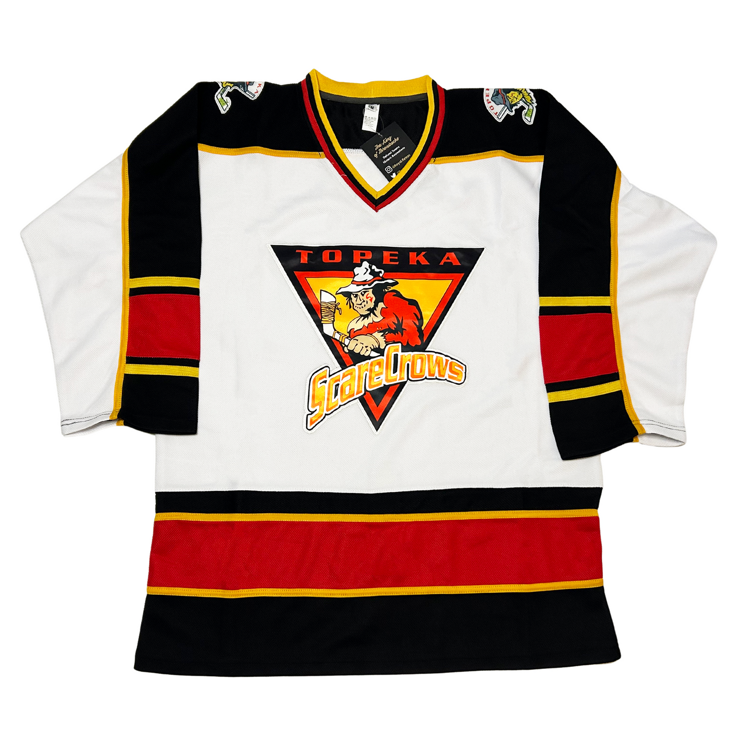Topeka Scarecrows hockey jersey white. Black shoulders. Half white sleeves, half black, separated by a gold line. Thin gold, thin black, thick red, thin black, thin gold ban mid-sleeve. Black, and red ban separated by gold line at waist. Logo in center is upside down black triangle. TOPEKA in red above a scarecrow holding a hockey stick over a gold triangle outlined in red. Stylized SCARECROWS in gold arched across bottom of main Triangle. Royal Retros