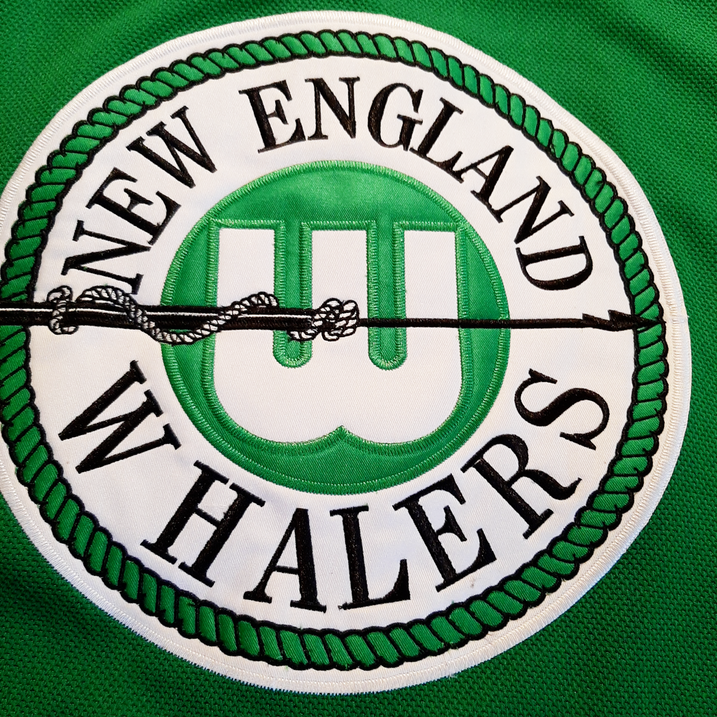 1972 New England Whalers Jersey close-up logo Circular logo with white outline. New England above in circle, Whalers below. W on green background in center of circle with black harpoon across Royal Retros