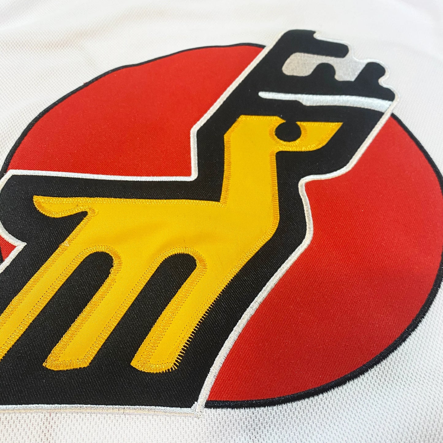 Michigan Stags Jersey close-up logo of gold deer outlined in black with black antlers over red circle on white jersey Royal Retros