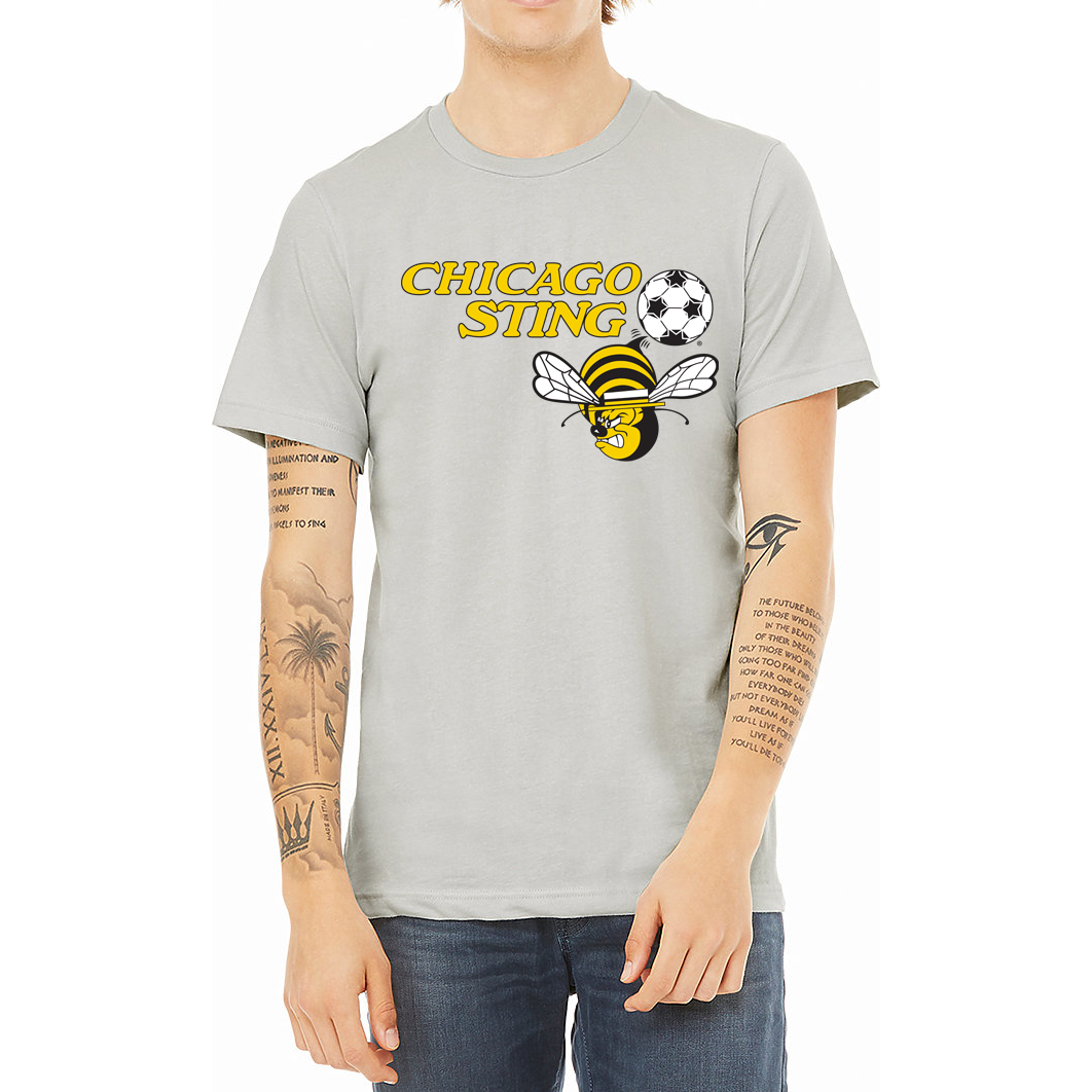 Chicago Sting T-shirt silver. "Chicago" slightly over "Sting" to the left in gold. Black and white soccer ball to the right with angry cartoon be with white wings below. Royal Retros. 