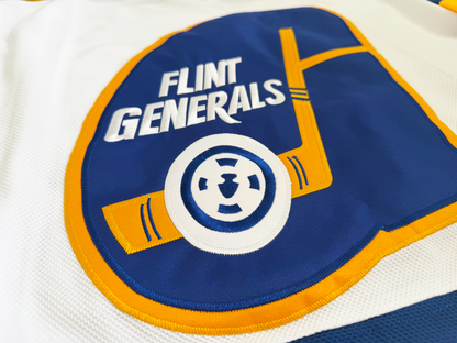 Close-up of the stitched Flint Generals crest on the white jersey — a blue teardrop G outlined in gold, FLINT GENERALS in white lettering, a gold hockey stick and a white puck with a blue wheel emblem