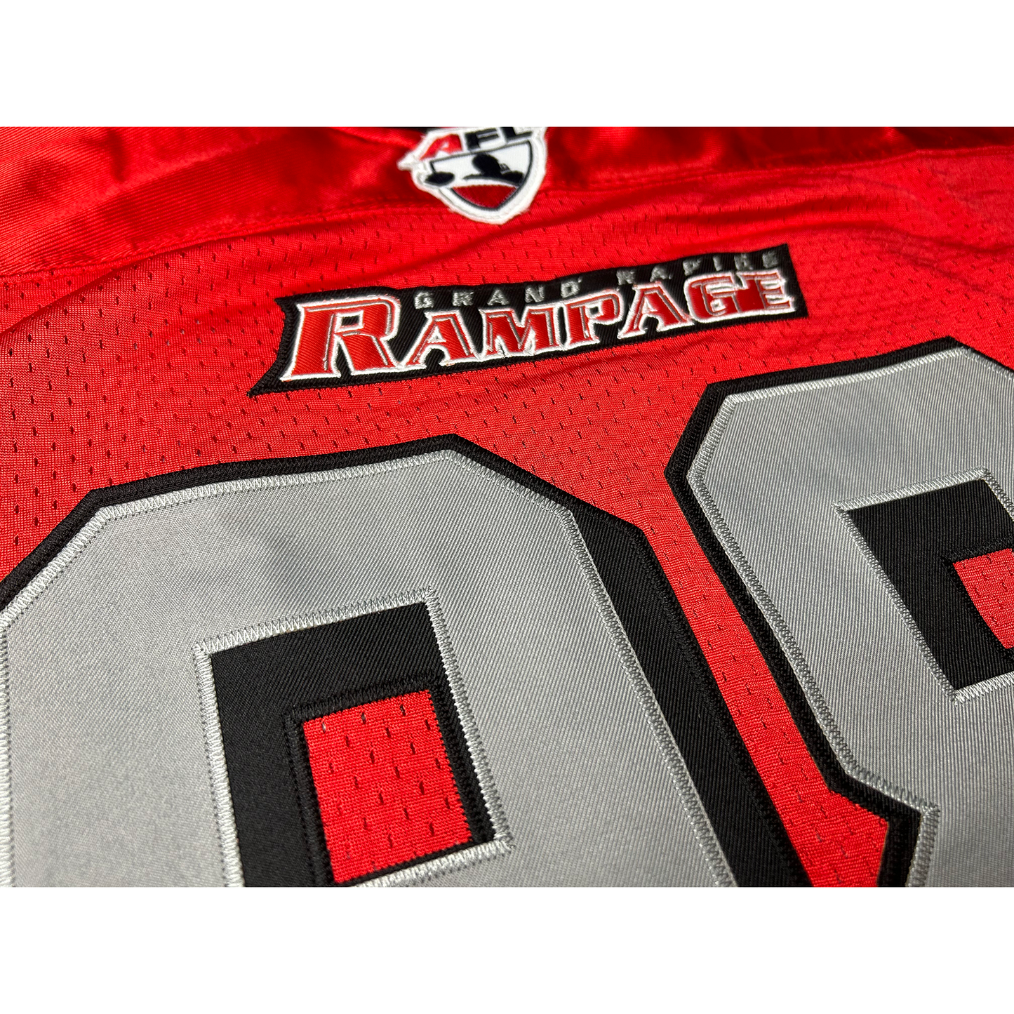 Grand Rapids Rampage Arena Football League jersey, close-up. Red. Black, red, black collar. AFL 2000s logo just below collar, small. Rampage logo of red charging rhino head on sleeves. Black, red, and black trim at sleeve ends. #98 in silver with black shadow outline large on chest, small on sleeves. Royal Retros