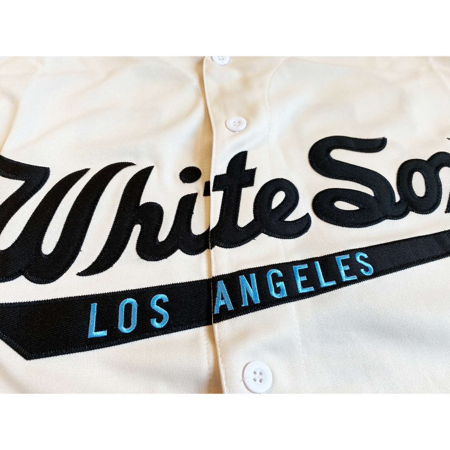 Close up of Los Angeles White Sox negro league button-up jersey white. Dark blue, light blue, dark blue trim on sleeve ends. White Sox script in blue across chest with Los Angeles in light blue on flare banner underneath. Interlocking LA on dark blue diamond with light blue trim. Royal Retros