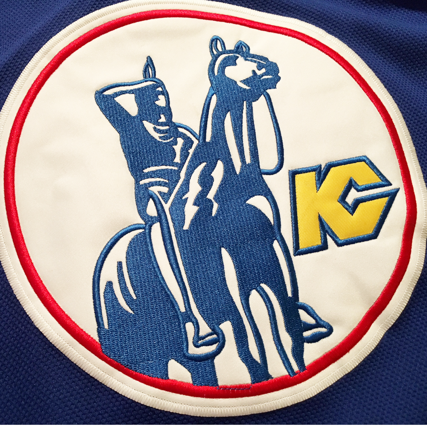 Kansas City Scouts Jersey Close-up of logo crest. Native American on horseback in blue on white background in red circle. Stylized KC in gold with blue trim.Royal Retros